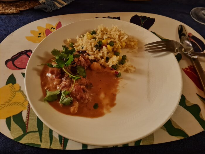 Tallrik med fiskgryta av torsk, sötpotatis och kokosmjölk med koriander, samt basmatiris blandat med paprika, majs och ärtor på en blommig duk.