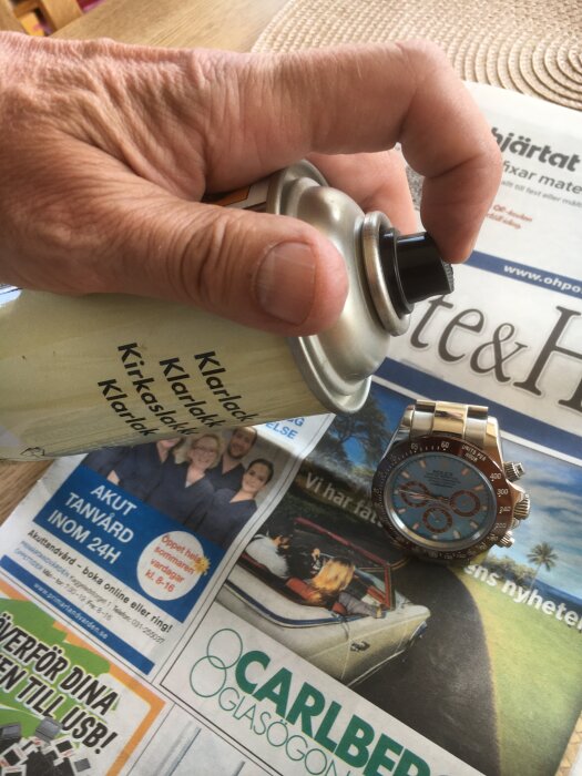 Hand håller i sprayburk över tidning och klocka.