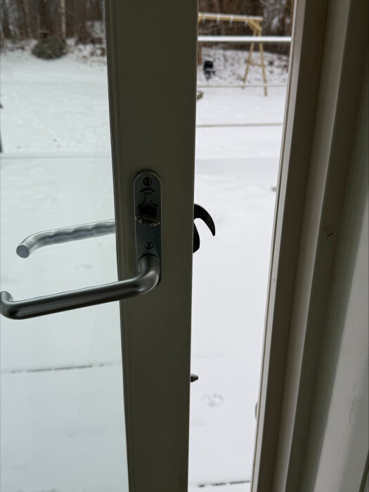 Altandörr med ett defekt lås, handtaget är sladdrigt och dörren stänger inte helt, vilket syns i springan vid dörrens kant. Snötäckt trädgård i bakgrunden.