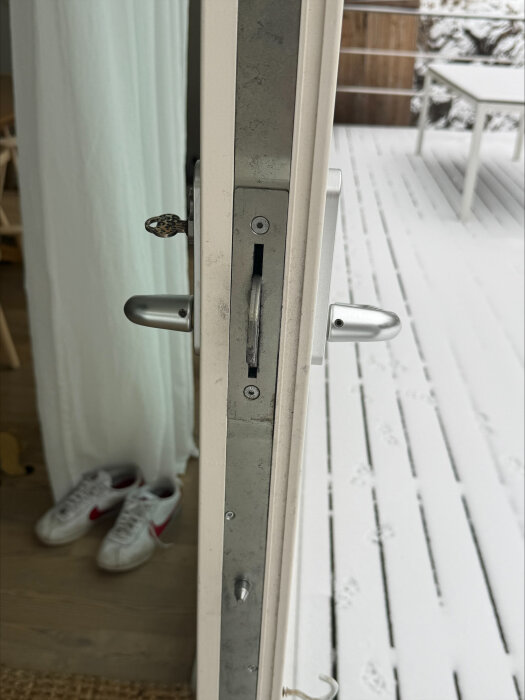 Ytterdörr med lås sett från sidan, dörren står öppen mot en snötäckt altan. Ett par vita sneakers syns inomhus.