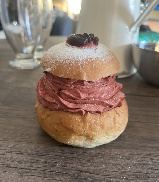 Bulle med florsocker och rödgrädde fyllning, toppad med mörk chokladbit på ett träbord.