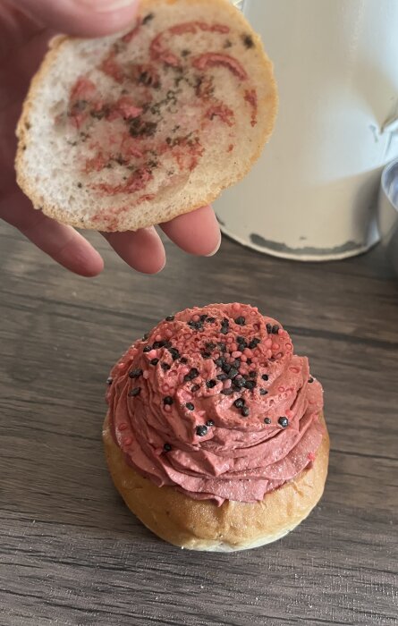 En öppnad semla med rosa grädde och svarta strösselkulor på ett bord. En hand håller semlans lock ovanför.