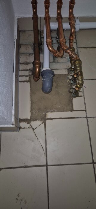 Vägg med flera kopparrör och ett vitt avloppsrör som går genom golvplattor med betong runt.