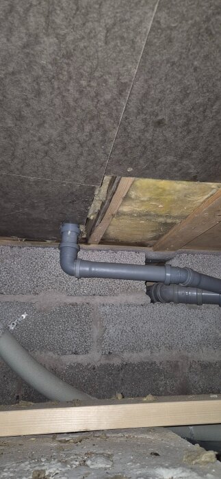 Grå rörinstallation i en vägg med isolering och betongblock, omgivna av träreglar och byggmaterial.