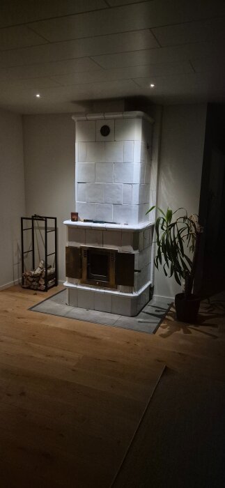 Effektbelyst kakelugn med vit kakel i ett rum, omgiven av ett vedställ och en växt i kruka.