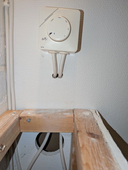 Temperaturregulator på badrumsvägg med synliga kablar genom hål, träbalkar och ett hål i väggen för kablar.