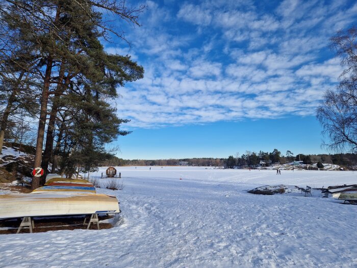 Snötäckt sjö med båtar längs strandkanten, omgiven av tallar och människor som promenerar i vinterlandskapet under en blå himmel med moln.