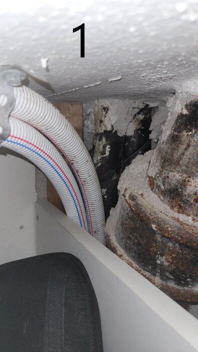 PEX-rör med röd och blå markering, omgivna av äldre rör och skadad vägg. Bild ovanifrån, visar avloppsledningens dragning i huset.