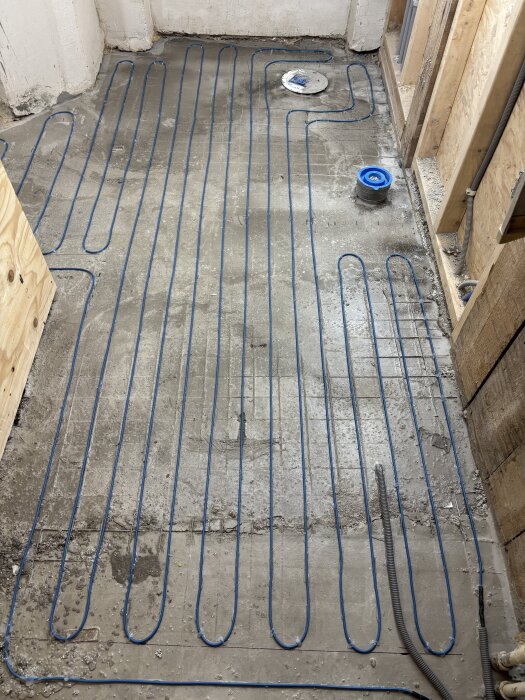 Golvvärmeslingor installerade på betonggolv i ett rum under renovering, med väggar av träreglar och byggmaterial.