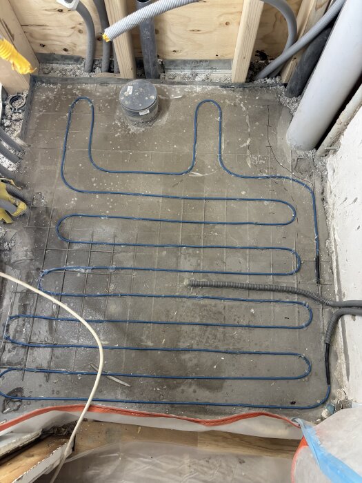 Golv med nylagd golvvärmeslinga i blått, omgiven av träreglar och installationsrör, under konstruktion.