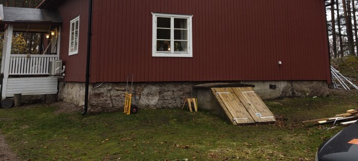 Rött trädhus med vit veranda, fönster, källaringång med träportar, och en gul pirra på gräsmatta. Skog i bakgrunden.