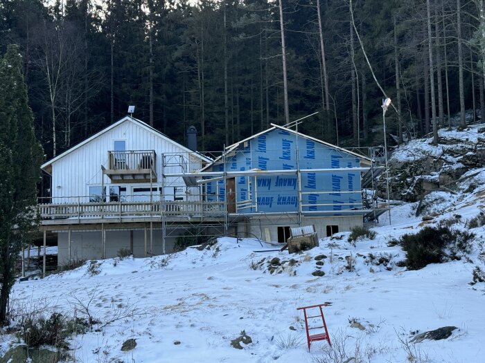 Två snöklädda hus vid skogskanten, ett under renovering med byggställningar och blått täckmaterial. Snön täcker marken.