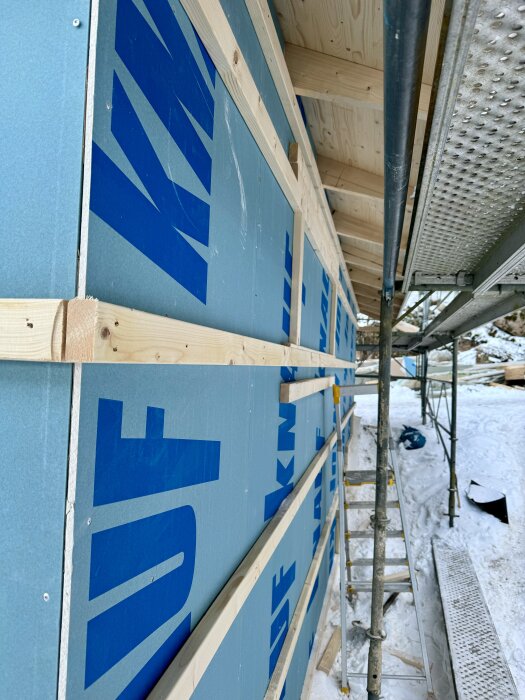 Byggnadsställning vid fasad med isoleingsskivor, snö och verktyg i bakgrunden en kall vinterdag.