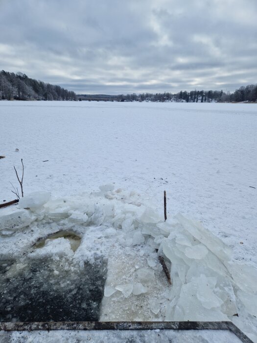 Isvaksöppning i en frusen sjö med snötäckt landskap och skog i bakgrunden under molnig himmel.