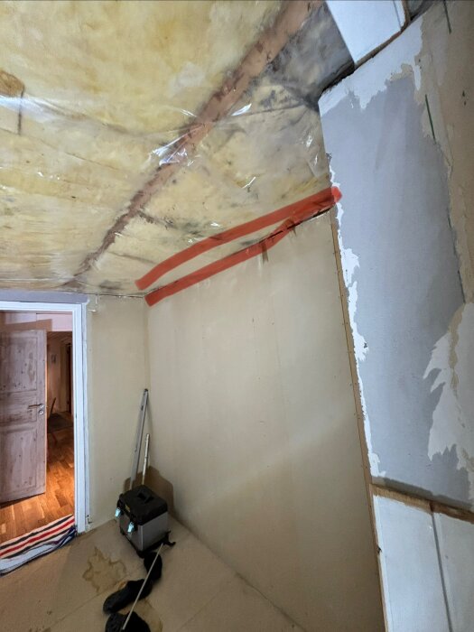Rum under renovering med synlig isolering i taket och orange tejp. Stege, verktygslåda och halvfärdig väggbehandling.