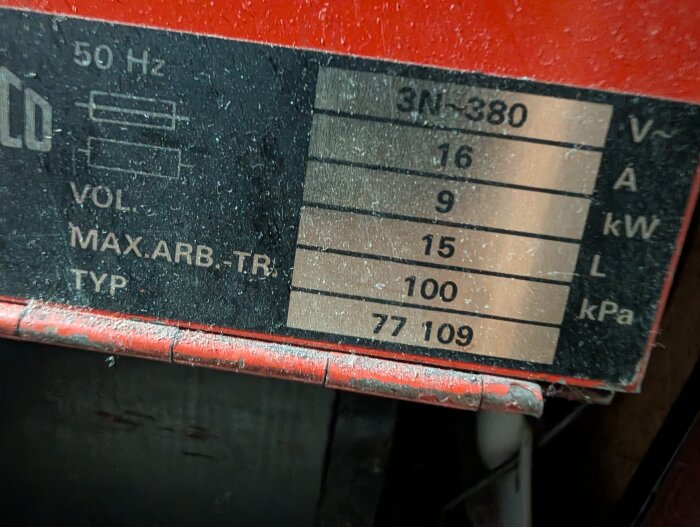 Vit metallskylt med tekniska specifikationer, inklusive 9 kW och 16 A, på röd bakgrund.