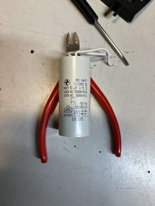 Motorkondensator 5 µF för cirkulationspump tillsammans med röd avbitartång och skruvmejsel på arbetsbord.