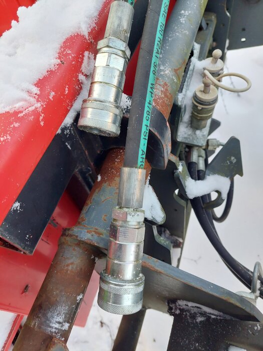 Hydraulkopplingar monterade på en snöplog med rostiga metalldelar och snötäckta ytor, fokus på de metalliska kopplingarna.