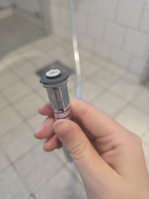Hand som håller en silverfärgad ventilkomponent i ett kaklat badrum.