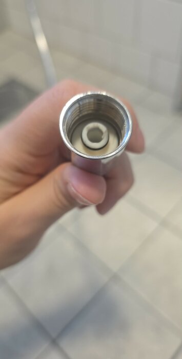En hand håller i en metallmutter med invändig gänga, antagligen en del av en VVS-installation, mot en suddig bakgrund av kakelgolv.