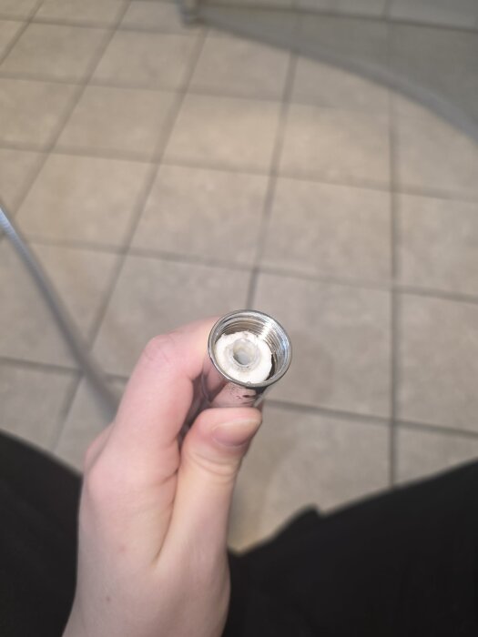 Hand håller en duschslang i metall, öppet ände uppåt med vit plastdel inuti, över ett golv med kakelplattor.