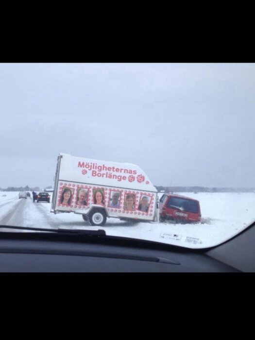 Släpvagn i snö vid vägkant, dekorerad med porträtt och texten "Möjligheternas Borlänge", kopplad till röd bil.
