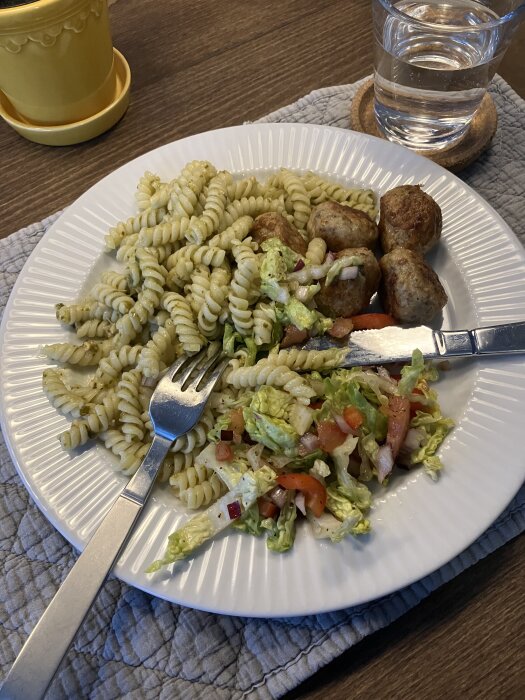Tallrik med spiralmakaroner med grön pesto, italienska kycklingbullar och sallad, bredvid glas med vatten.