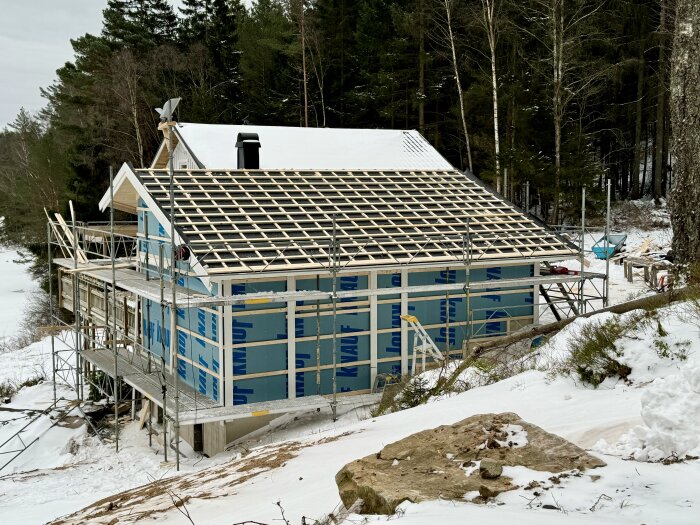 Byggarbetsplats i vinterskog med halvfärdigt tak och ställningar runt huset för renovering och plåtslageriarbete.