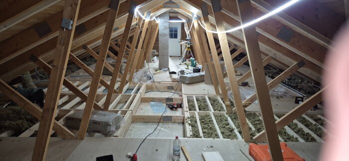 Byggprojekt på vinden med träreglar, isolering och öppningar där spånplattor ska läggas. Trappa kommer att placeras här.