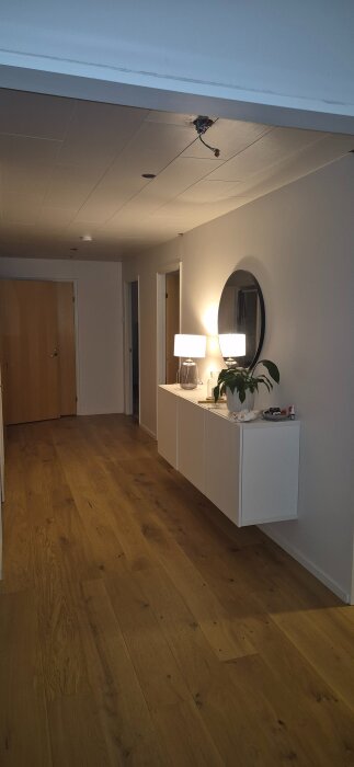 Hall med trägolv, väggskåp med lampa och växt, dörrar, och plats för kommande trappa.
