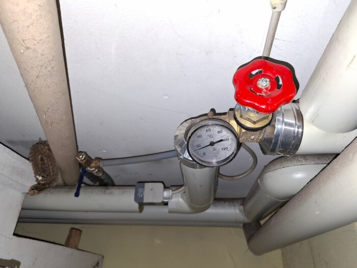 Rörsystem i källare med en termometer och röd ventil, del av vedpannans värmesystem.