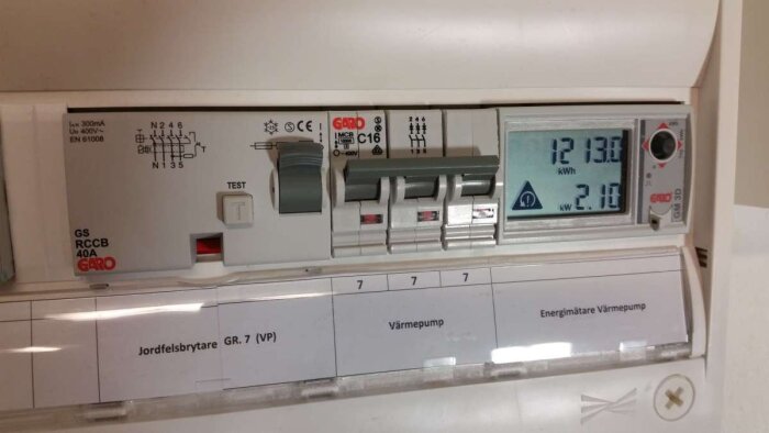 Elmätare för värmepump monterad på en DIN-skena, visande energiförbrukning i kWh och kW med jordfelsbrytare till vänster.