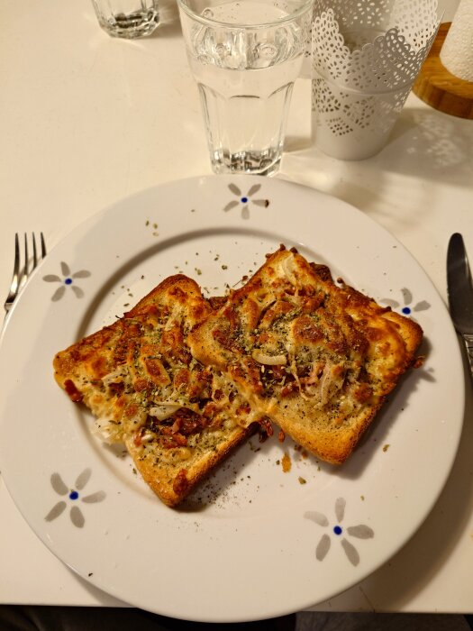 Två skivor rostat bröd med smält ost och kryddor på en tallrik med blommönster, serverad med ett glas vatten.