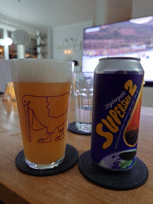 Ölglas med en gul dryck och en burk från Stigbergets på ett bord, bakgrund med TV som visar sport.