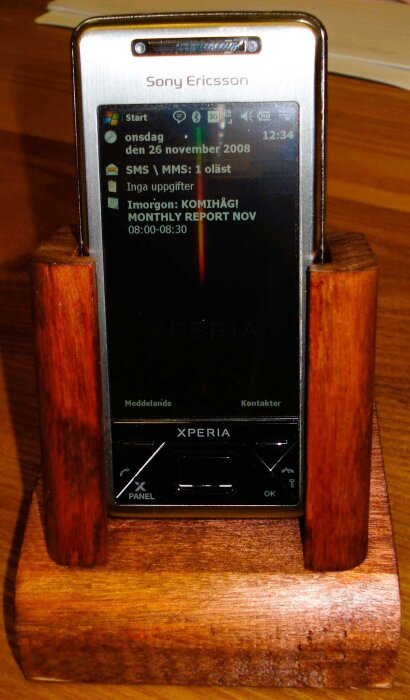 En äldre Sony Ericsson mobil i ett hemmagjort trähållare, skärmen visar datumet 26 november 2008.