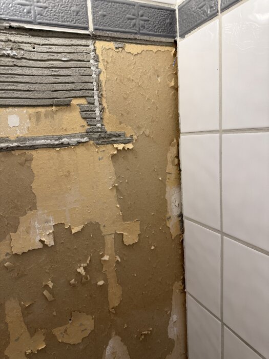 Vägg med gammalt, delvis bortskalat gips och intilliggande vita kakelplattor i ett badrum.