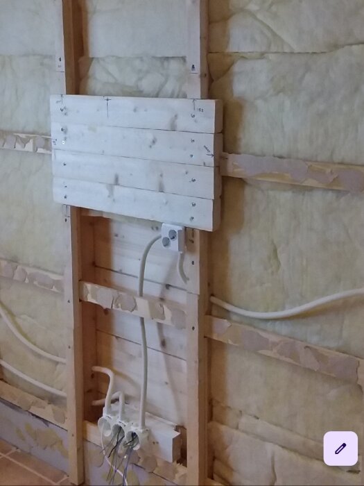 Vägg under konstruktion med trästolpar, isolering och elledningar dragna för installation.