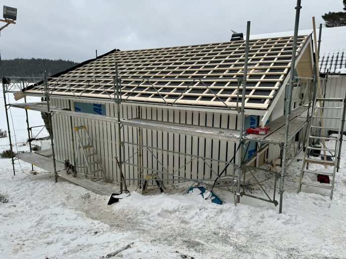 Bygget av en byggnad med takstolar under uppförande, täckt av ställningar, omgiven av snö, i en kall miljö.