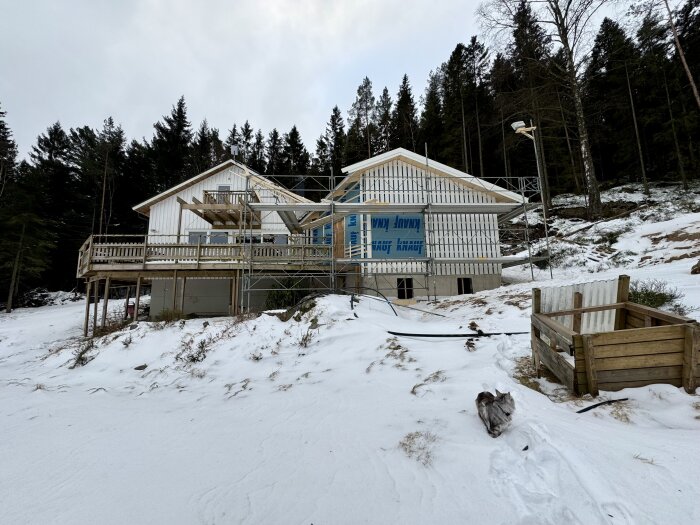 Hus under renovering med byggnadsställning och snö på marken, skog i bakgrunden. En grå katt går i snön framför huset.