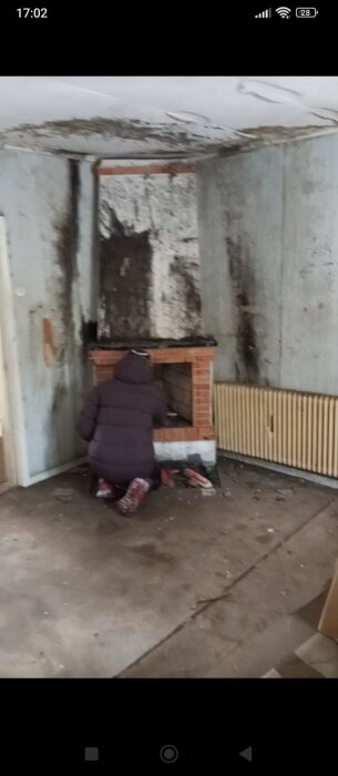 Person sitter framför en gammal braskamin i ett slitet rum. Väggarna är sotiga och en radiator syns bredvid kaminen.