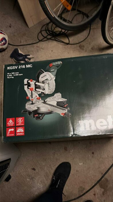 Kartong med kap- och gersåg av märket Metabo KGS 216 MC, fotograferad på ett garagegolv med cykelhjul i bakgrunden.