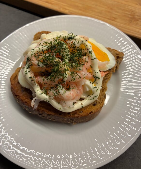 Räksmörgås på en tallrik med rågbröd, ägg, majonnäs och dill på toppen.