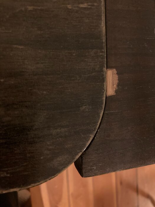 Skada i fanér på ett mörkt trämöbelstycke, där en del av det underliggande materialet är synligt.