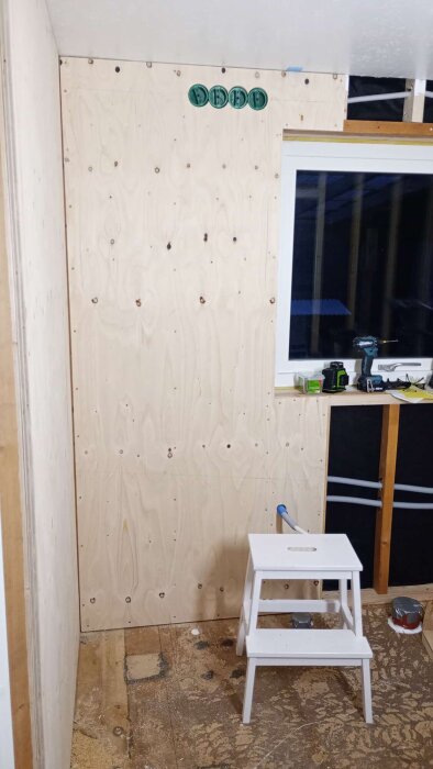 Vägg täckt med plywood i renoverat badrum, med flera eldosor högst upp. En vit stege står på golvet framför väggen. Verktyg syns i fönsterkarmen.
