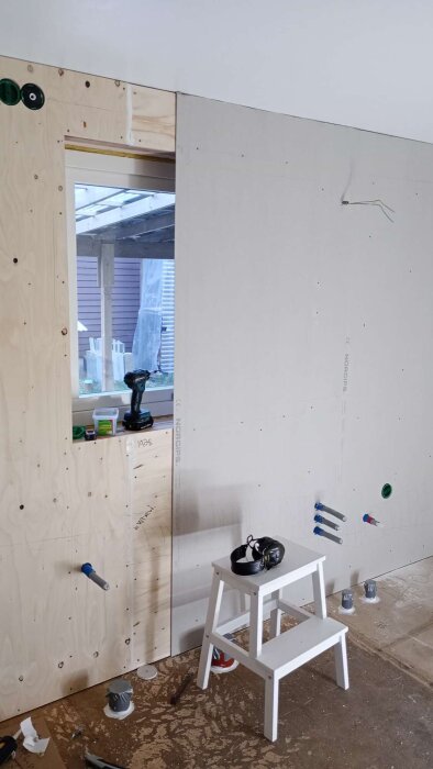 Renovering med plywood och gipsväggar, fönsteröppning och verktyg synliga; stege framför vägg med eluttag och rör.