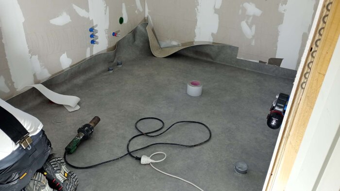 Badrum under renovering med plastmatta som läggs, verktyg på golvet och synliga rör på väggen.