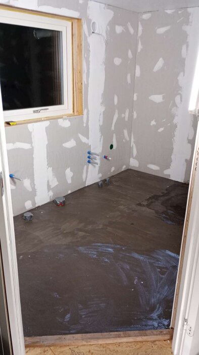 Badrum under renovering med gipsväggar och förberett golv. Flera vattenanslutningar i väggen och ett fönster utan lister.