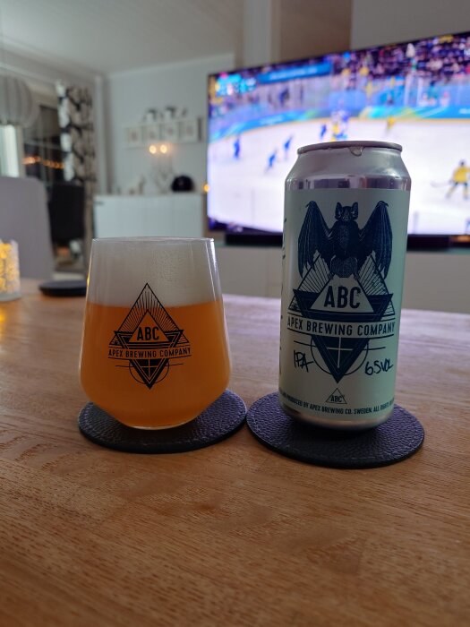 Ett glas och en burk Apex Brewing Company IPA på ett bord, med en ishockeymatch på TV i bakgrunden.