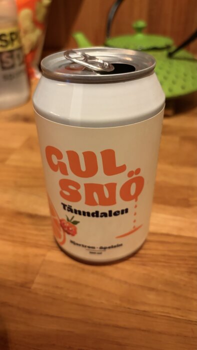 En öppnad läskburk med etiketten "Gul Snö" på ett träbord, med en grön tekanna i bakgrunden.