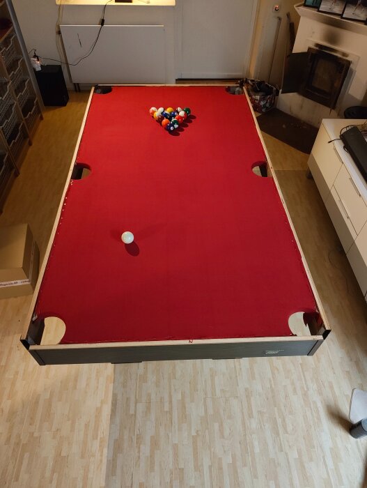 Biljardbord med röd filt, placerad i ett vardagsrum. Poolbollar står uppradade i triangel och en vit köboll ligger redo.